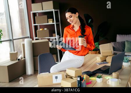 Elegant businesswoman in vibrant workspace checking documents and holding coffee cup with parcels around her workspace table Stock Photo