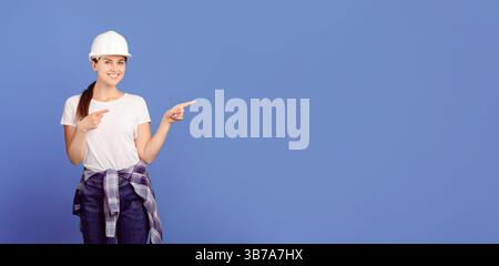 Smiling handyworkers couple wearing casual clothes and protective hard hats pointing aside Stock Photo