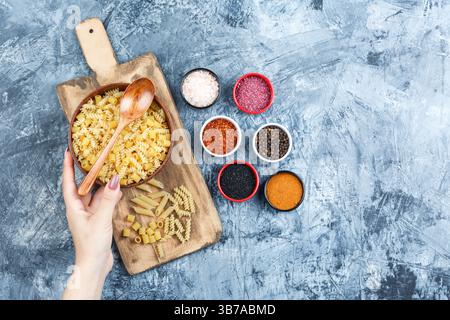 Some hand holding bowl of pasta with spices, wooden spoon on grey plaster and cutting board background, top view. Stock Photo