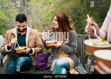 Young couple having lunch outdoors on a park bench, sharing a relaxed and friendly moment in the city. Stock Photo