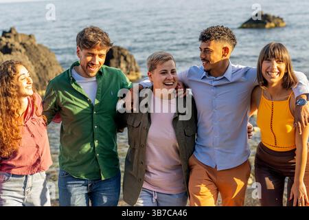Multiracial group of young friends hugging and laughing on a rocky beach at sunset, enjoying summer together. Stock Photo