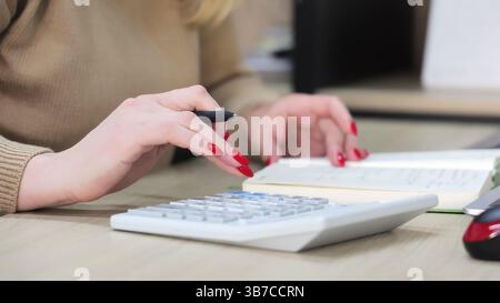 Close-Up of Woman Using Calculator and Writing Notes. Close-up of a womans hands with red nails using a calculator and writing notes, focusing on Stock Photo