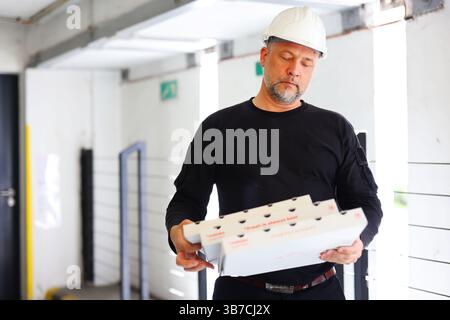 Construction worker in hard hat holding pizza boxes, taking a break at the worksite Stock Photo