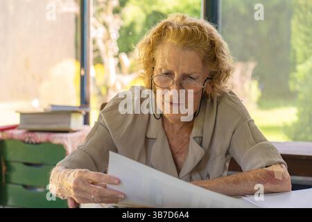 Focused elderly woman wearing glasses reading important documents while sitting at her home office desk, concentrating on paperwork and administrative Stock Photo