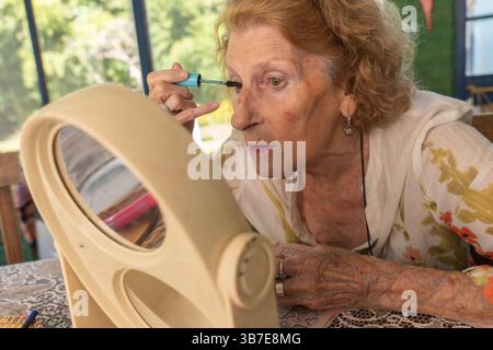 Elderly woman applying mascara with care, using a small mirror on the table, enhancing her beauty routine with precision and focus, embodying self-car Stock Photo