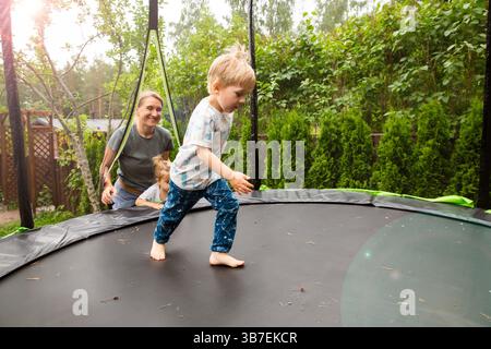 Happy mother playing with her children on a trampoline in the backyard on a sunny day. Family outdoor activity, summer fun and quality time with kids. Stock Photo