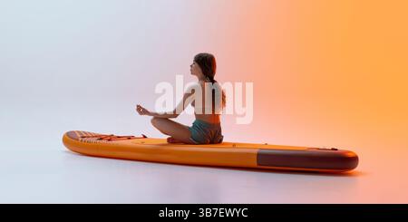 Calm young woman sits in meditative lotus pose on sup board in minimal indoor setup with gentle light gradient creating peaceful scene. Stock Photo