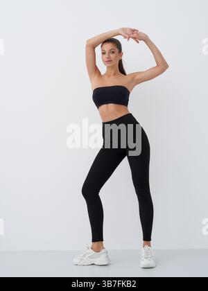 Woman posing confidently in black sportswear, including a strapless top and high-waisted leggings. She stands on one leg with arms gracefully arched Stock Photo