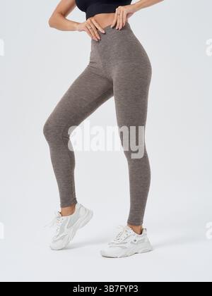 Athletic woman in gray leggings and white sneakers on plain background stands in a casual pose with her hand on waist, showcasing activewear and Stock Photo