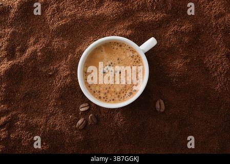 White ceramic coffee mug filled with foamy coffee in the center on a background of ground coffee and beans around. Top view. Stock Photo