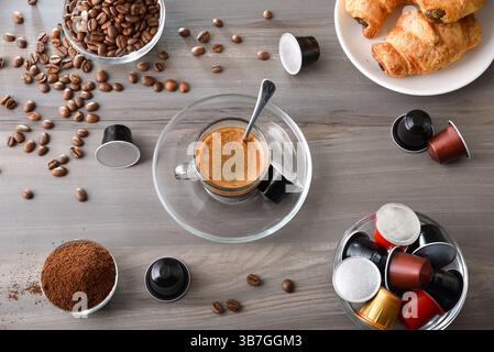 Breakfast with a cup of espresso in a glass and a saucer on a wooden table with bowls of coffee beans, ground coffee, capsules, and pastries. Top view Stock Photo
