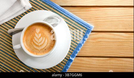 Foamy coffee with a flower pattern in a white ceramic cup and saucer on a bamboo tablecloth on a wooden table. Top view. Stock Photo