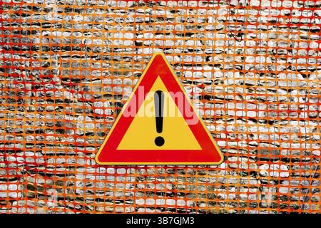 An exclamation point on a yellow background, in a red triangular sign. The texture of the yellow plastic mesh fence on the construction site. Stock Photo