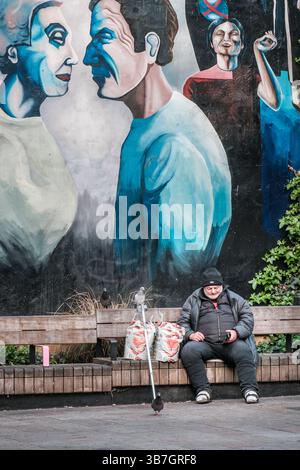 London, England - 28th April 2025: A homeless man on a park bench with pigeons checks his phone below a large painted mural at Whitfield Garden off To Stock Photo