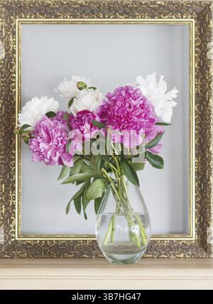 Pink and white peonies (Paeonia officinalis) in a vase in front of an empty picture frame on a sideboard, Siegen Stock Photo