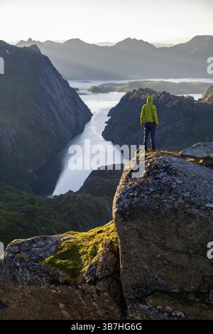 Mountaineer, young man standing on rocks and looking into the distance, at sunrise, fjord Trollfjord and mountains, hike to Trollfjord Hytta, at Troll Stock Photo