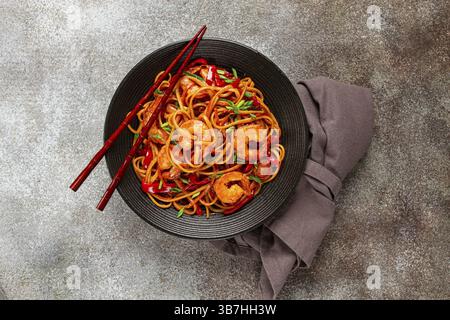Udon stir-fry shrimp noodles with red bell pepper and green onion, served in a black bowl, with chopsticks, gray background Stock Photo