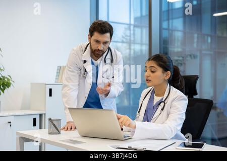 Two doctors collaborate in a modern office setting, focusing on a laptop while discussing medical information and data. The scene portrays professional teamwork. Stock Photo