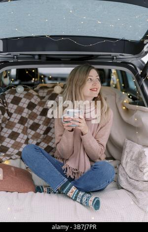 Smiling young woman with coffee cup sitting in car trunk and looking away. Stock Photo