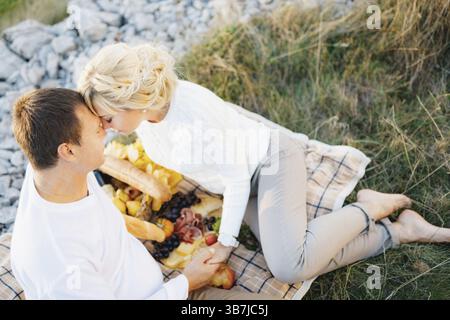 Barefoot woman leaned her forehead against man sitting on a blanket. Man and woman are holding hands. There is food on the blanket. Stock Photo