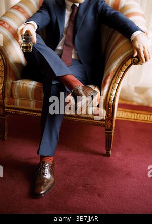Legs of a man in blue pants, red socks and brown shoes sitting in the chair holding glass in one hand. Stock Photo