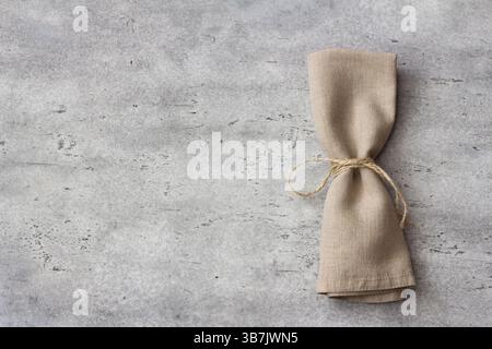 Top view of folded linen napkin tied with rope on gray textured stone surface. Minimal rustic table setting for food styling, eco-friendly design, or Stock Photo