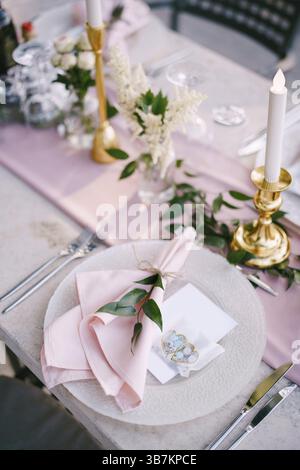 Wedding dinner table reception. Stone table with pink runner, plates with pattern and rag pink napkins, candles with led-light bulbs in the rays of su Stock Photo