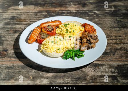 Big breakfast with bread, scrambled egg, sausages, roasted tomato, baked bean, sauted mushroom, and spinach on white plate. Isolated by wooden table Stock Photo