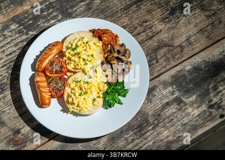 Big breakfast with bread, scrambled egg, sausages, roasted tomato, baked bean, sauted mushroom, and spinach on white plate. Isolated by wooden table Stock Photo