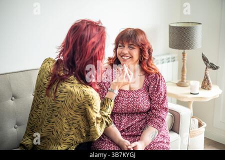 Two friends, a non-binary person and a curvy woman, sitting on a sofa, enjoying intimate moments of self-care while applying makeup and celebrating bo Stock Photo