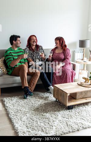 Three cheerful friends, including a non-binary person and a curvy woman, sitting on a sofa in a modern apartment, enjoying wine and laughter while cel Stock Photo