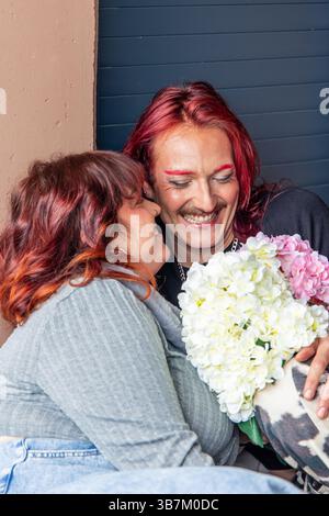 Gender fluid man with red hair and painted eyebrows embracing his curvy girlfriend holding a bouquet of hydrangeas, smiling and laughing, celebrating Stock Photo