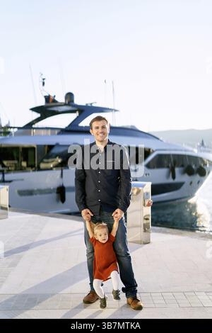 Man is holding his daughter by the hands while having a walk on a boat pier at sunset. Stock Photo