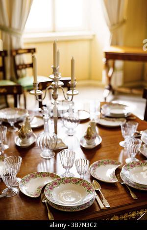 Festive table setting with clean empty plates, spoons, knives, wine glasses, decanters and a candlestick in the center on a wooden table in the room. Stock Photo