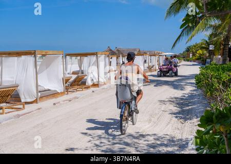 Isla Holbox, Mexico – 10 February 2025: A man rides his bicycle past beach cabanas and a golf cart taxi on a sandy path lined with palm trees and bout Stock Photo