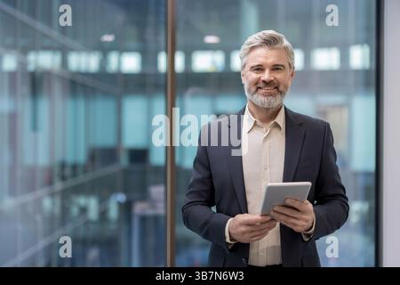 A smiling, distinguished businessman stands confidently holding a tablet in a modern office setting with large windows. Stock Photo