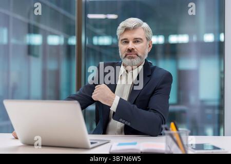 A serious-looking, mature businessman sits at a desk, staring at the camera with a laptop in front of him in an office setting. Stock Photo
