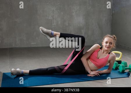 Young woman exercising with resistance band, doing side-lying leg lifts on yoga mat during home fitness workout. Stock Photo