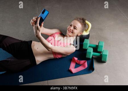 Happy woman taking selfie after home workout, lying on exercise mat with fitness equipment around her. Stock Photo