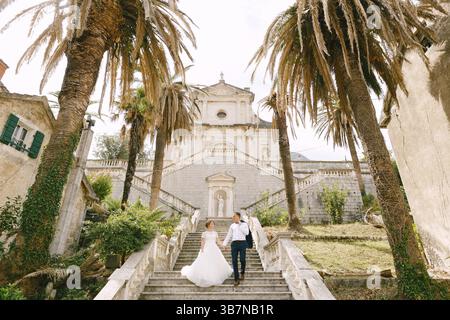 The bride and groom are walking down the stairs of the Nativity of the Blessed Virgin Mary church in Prcanj and holding hands. Stock Photo