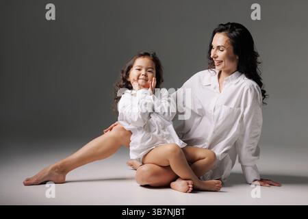 Joyful mother and daughter wearing oversized white shirts, sitting on studio floor, laughing and bonding together. Stock Photo