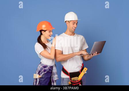 Young couple in hardhats using laptop during house repair, blue background Stock Photo