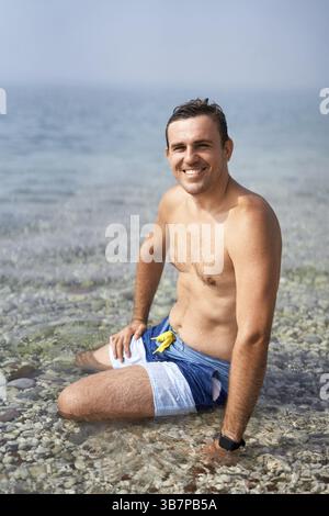 Young smiling man sitting in the water near the shore. Stock Photo
