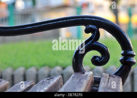 Close-up of decorative wrought iron armrest and aged wooden slats on a vintage park bench, with blurred grass and park background. Stock Photo