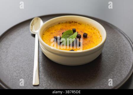A creamy creme brulee, topped with chocolate and a fresh mint leaf. A delicious dessert on a dark plate with a silver spoon, ready to be savored. Stock Photo