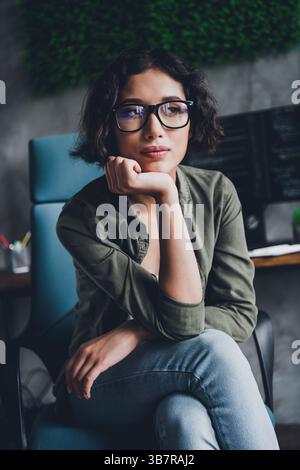 Young female programmer working remotely on a computer in a modern office, wearing casual attire, glasses, and feeling confident Stock Photo