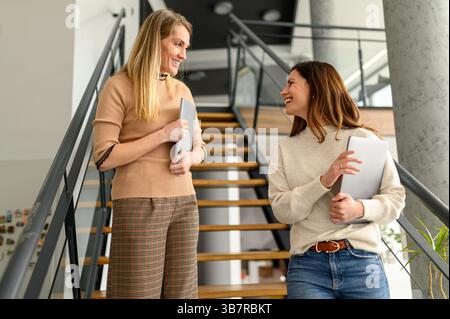 Low angle view of cheerful female colleagues with laptops talking while standing on steps in modern office Stock Photo