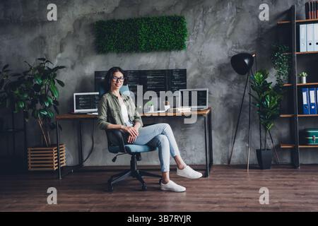 Young female programmer in casual attire sitting at a stylish home office with multiple screens displaying code Stock Photo