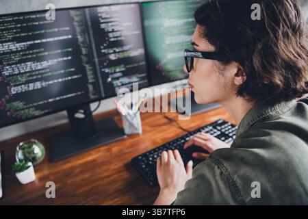 Young female programmer working on code at a modern workspace indoors using multiple screens, focused on software development Stock Photo