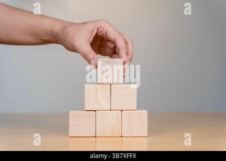 A hand places a wooden block atop a pyramid structure made of blocks, symbolizing strategy, planning, or construction in a professional setting, isola Stock Photo